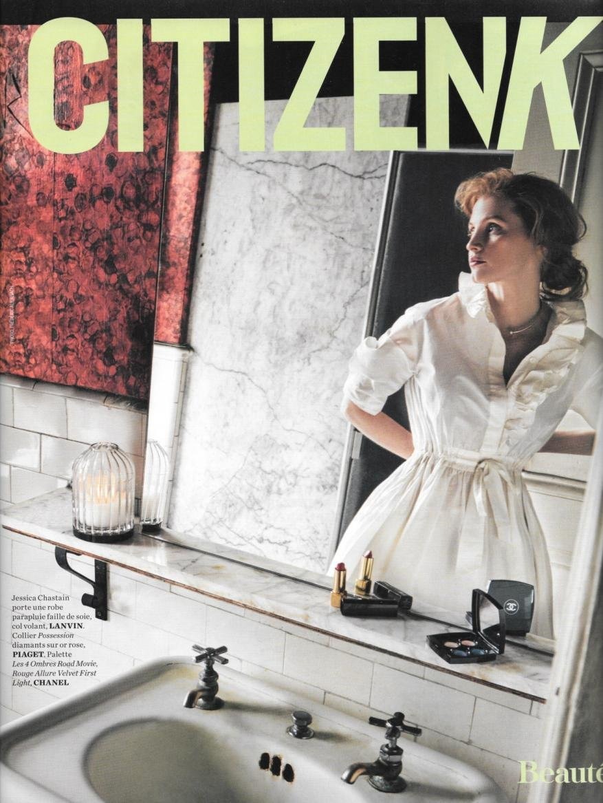 Magazine cover shot of a red-haired woman in a white dress reflected in a vintage bathroom mirror with beauty products and a sink.