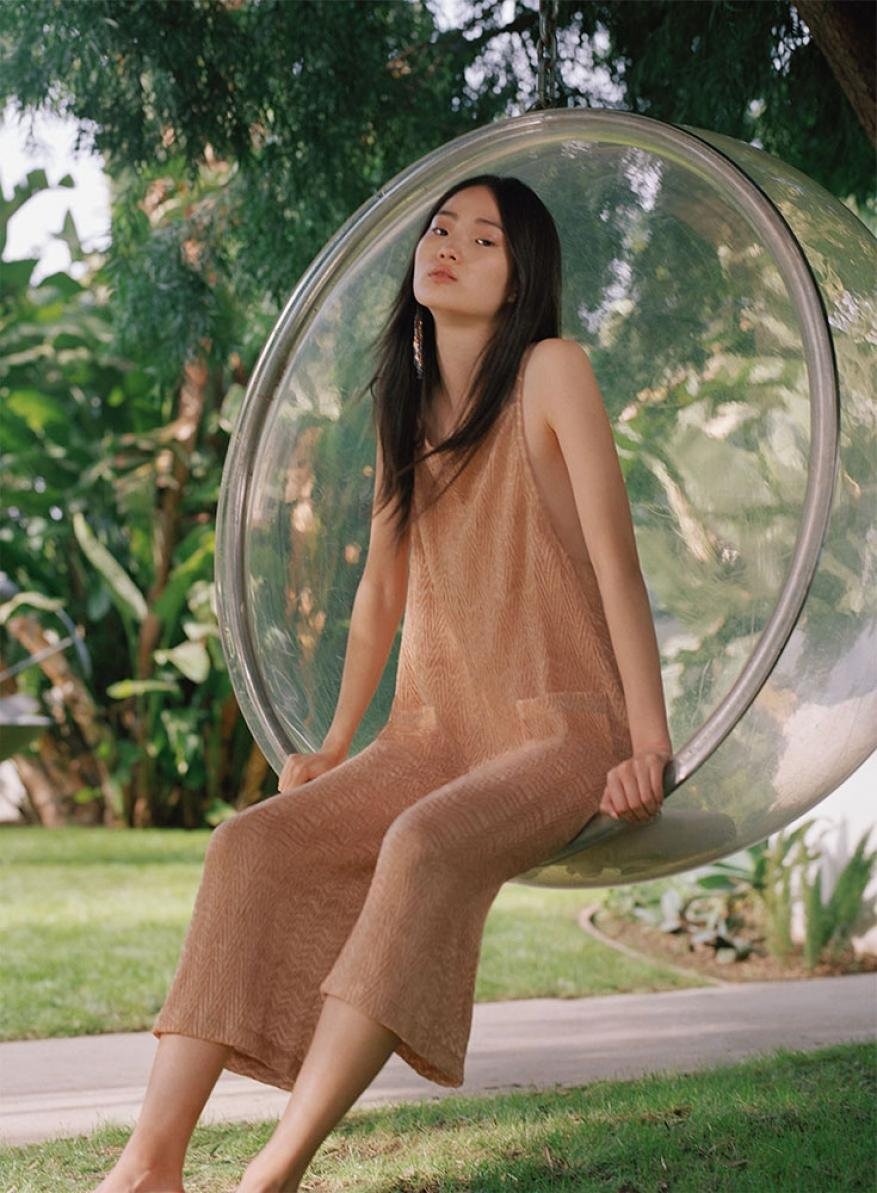 Young woman in a knit jumpsuit sitting inside a transparent bubble chair outdoors on a grassy lawn with trees in the background.