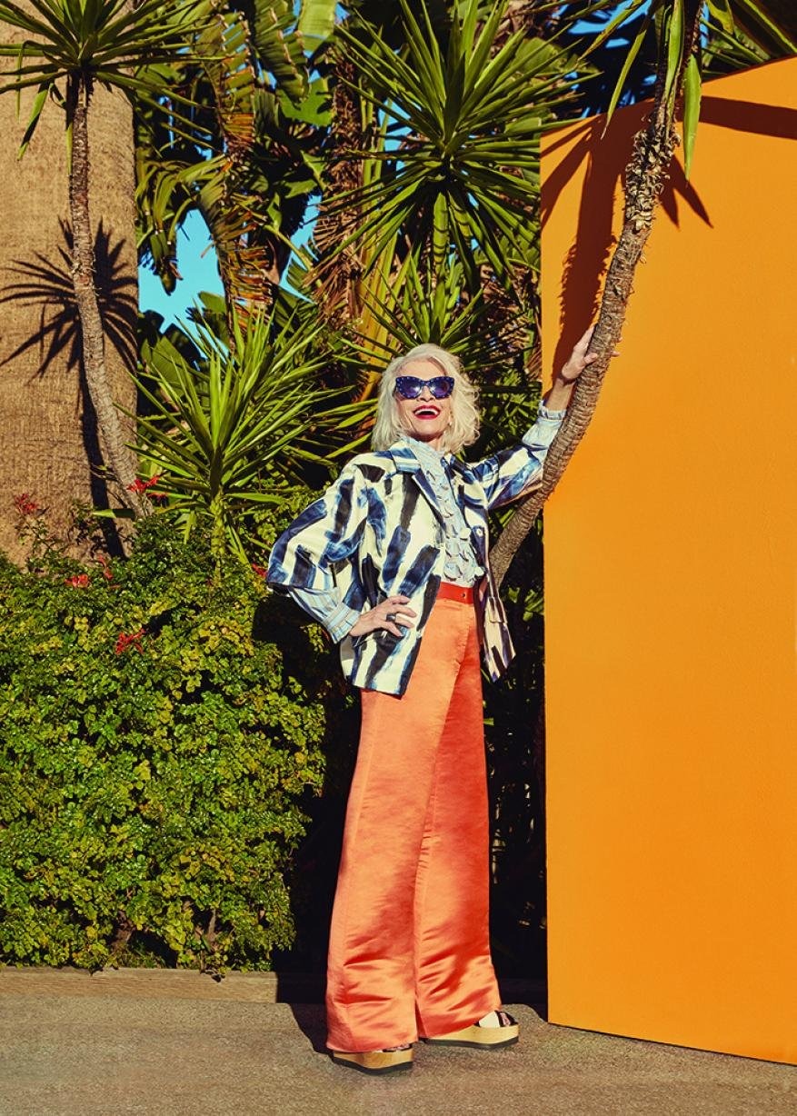 Fashionable senior woman with platinum blonde hair and sunglasses poses in orange palazzo pants and patterned jacket with palm trees.