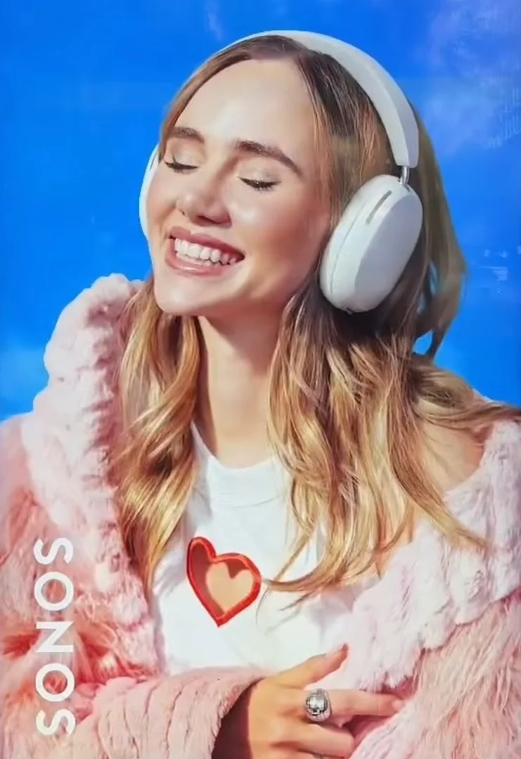 A smiling woman with blonde hair wears headphones and a pink fur jacket against a bright blue backdrop, with the Sonos brand logo visible.