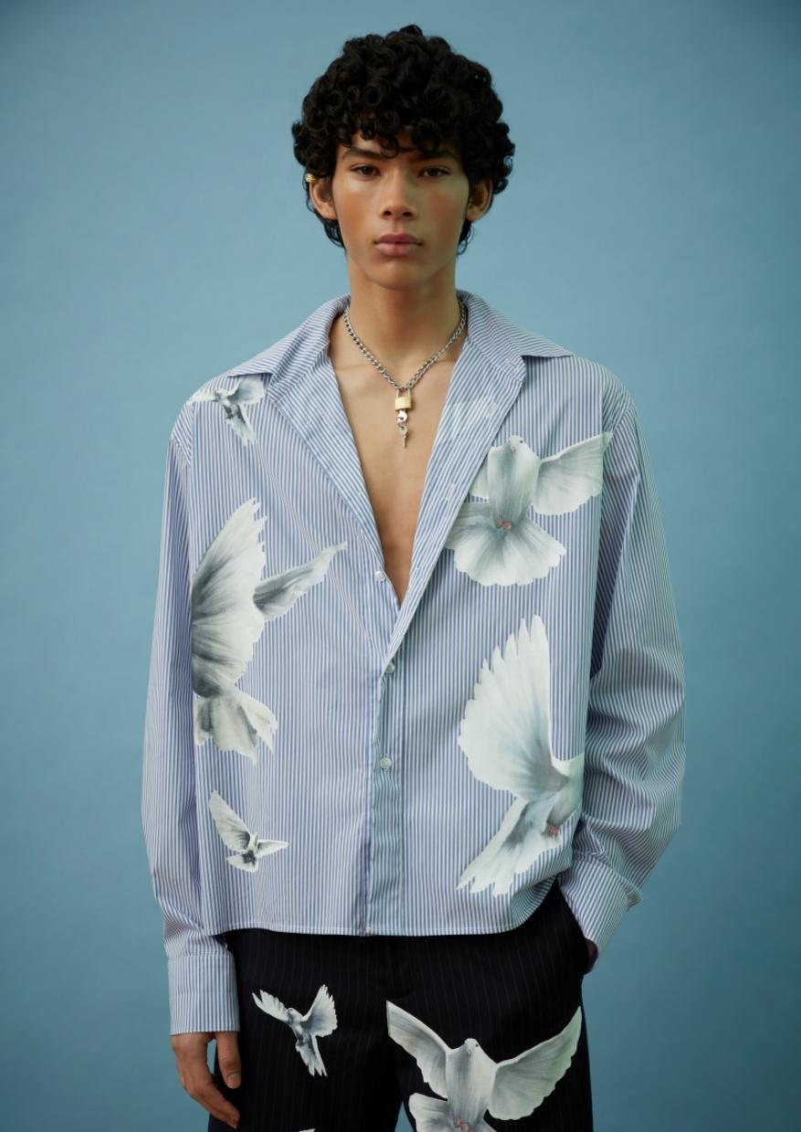 Young male fashion model poses in an unbuttoned striped shirt with dove prints against a light blue backdrop in a studio shot.