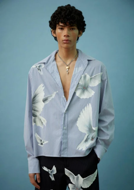 Young male fashion model poses in an unbuttoned striped shirt with dove prints against a light blue backdrop in a studio shot.