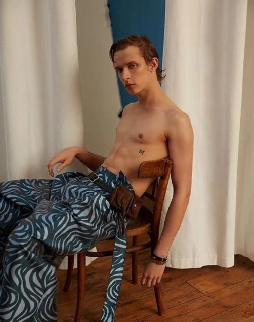 Young adult male poses shirtless in patterned pants, seated on a wooden chair against a simple studio backdrop with natural light.