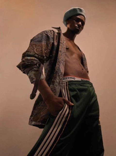 Stylish portrait of a Black male model wearing a blue beret, unbuttoned patterned shirt, Moschino briefs and green striped athletic pants.