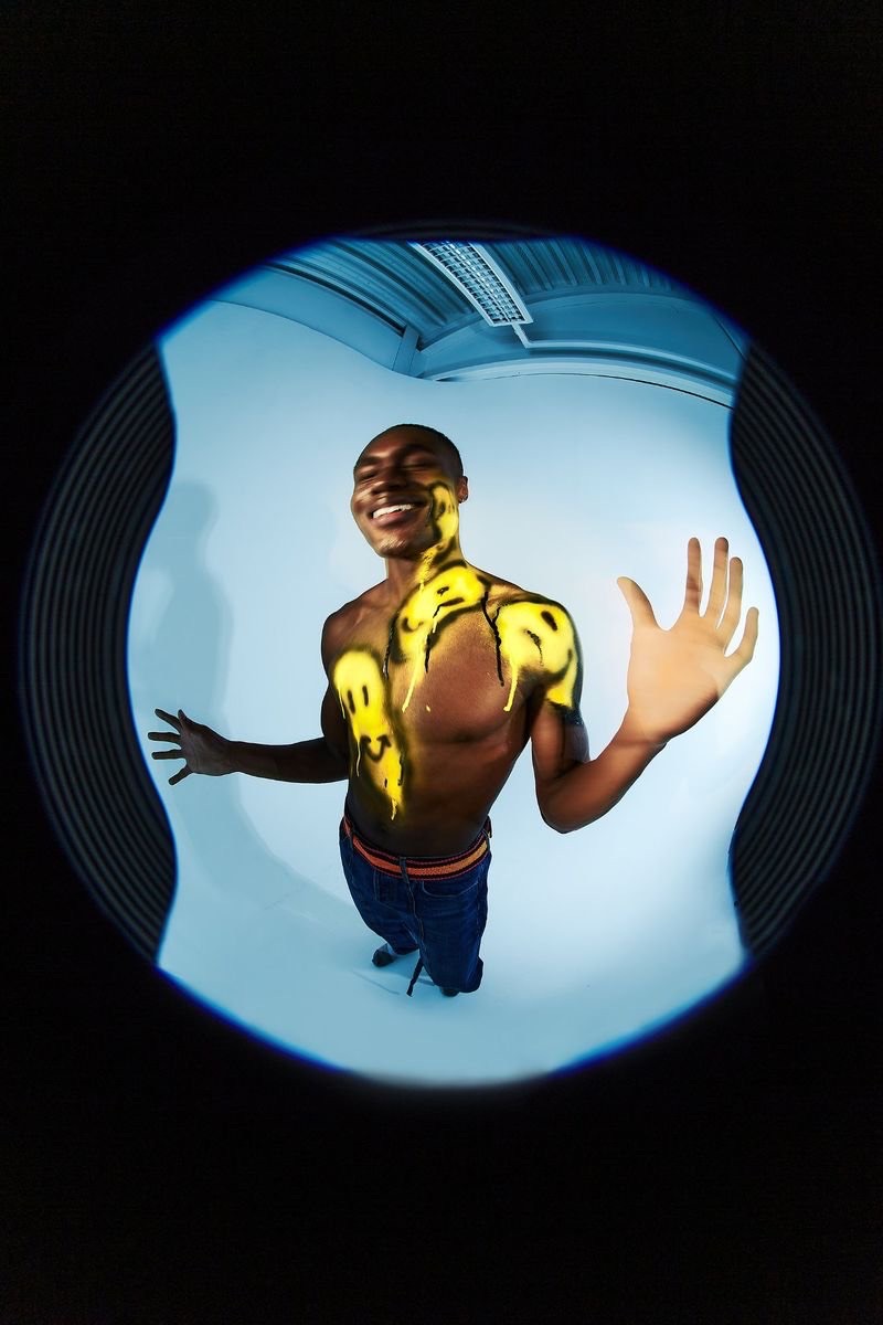 Full body shot of a smiling adult male with yellow smiley faces painted on his torso and arm, viewed through a circular fisheye lens.