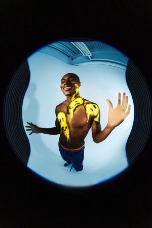 Full body shot of a smiling adult male with yellow smiley faces painted on his torso and arm, viewed through a circular fisheye lens.