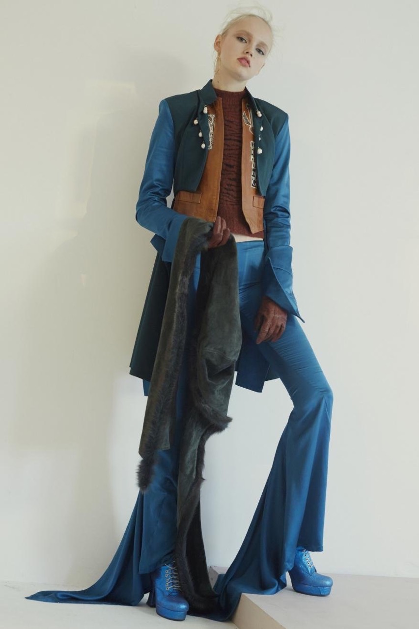 Full-length shot of a woman posing on a step wearing a blue pants suit, blue platform shoes, a dark shawl, and a brown top in a studio.
