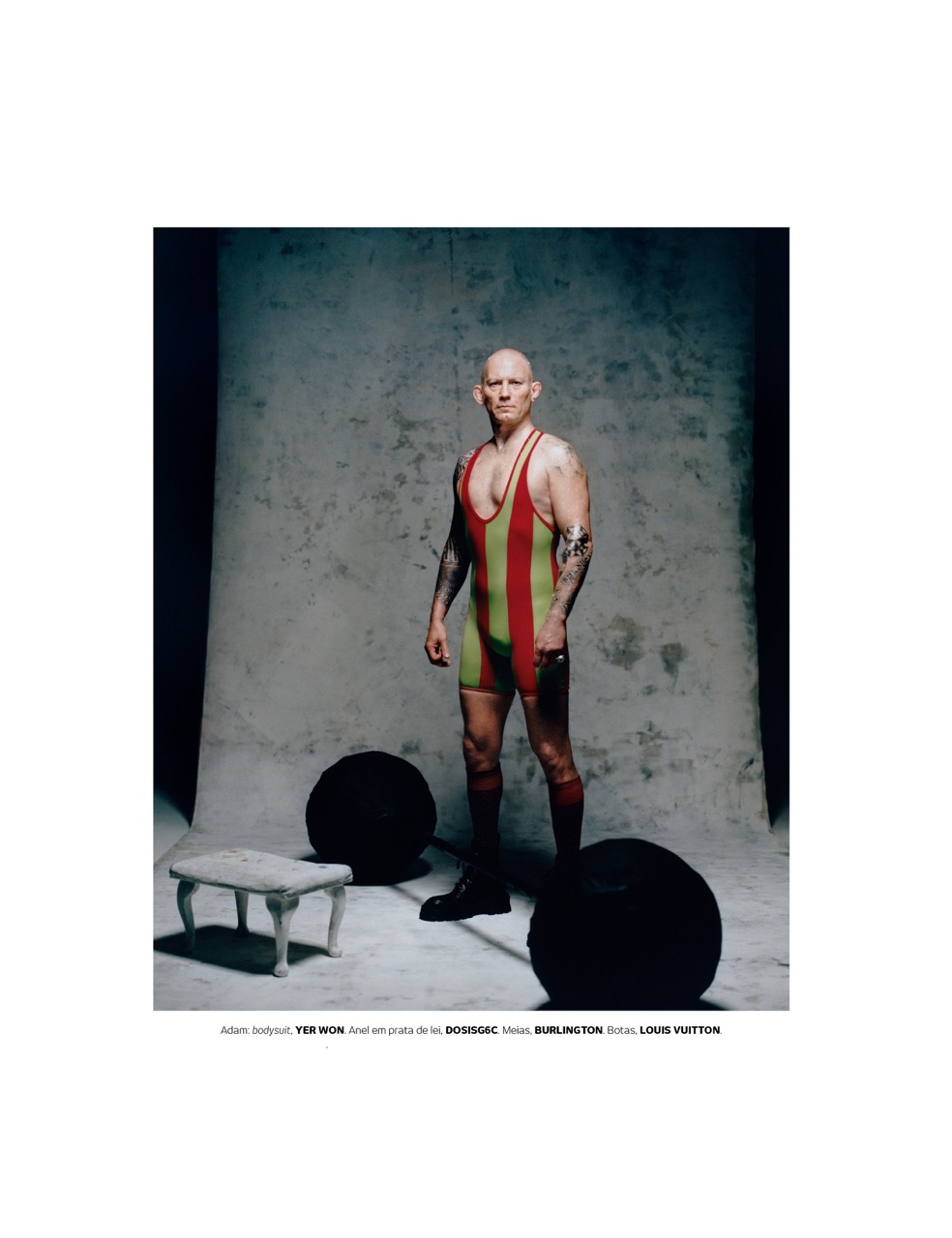A bald adult male stands in a vintage wrestling suit in a photo studio, flanked by two large black exercise balls and a small ornate stool.