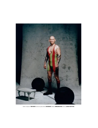 A bald adult male stands in a vintage wrestling suit in a photo studio, flanked by two large black exercise balls and a small ornate stool.