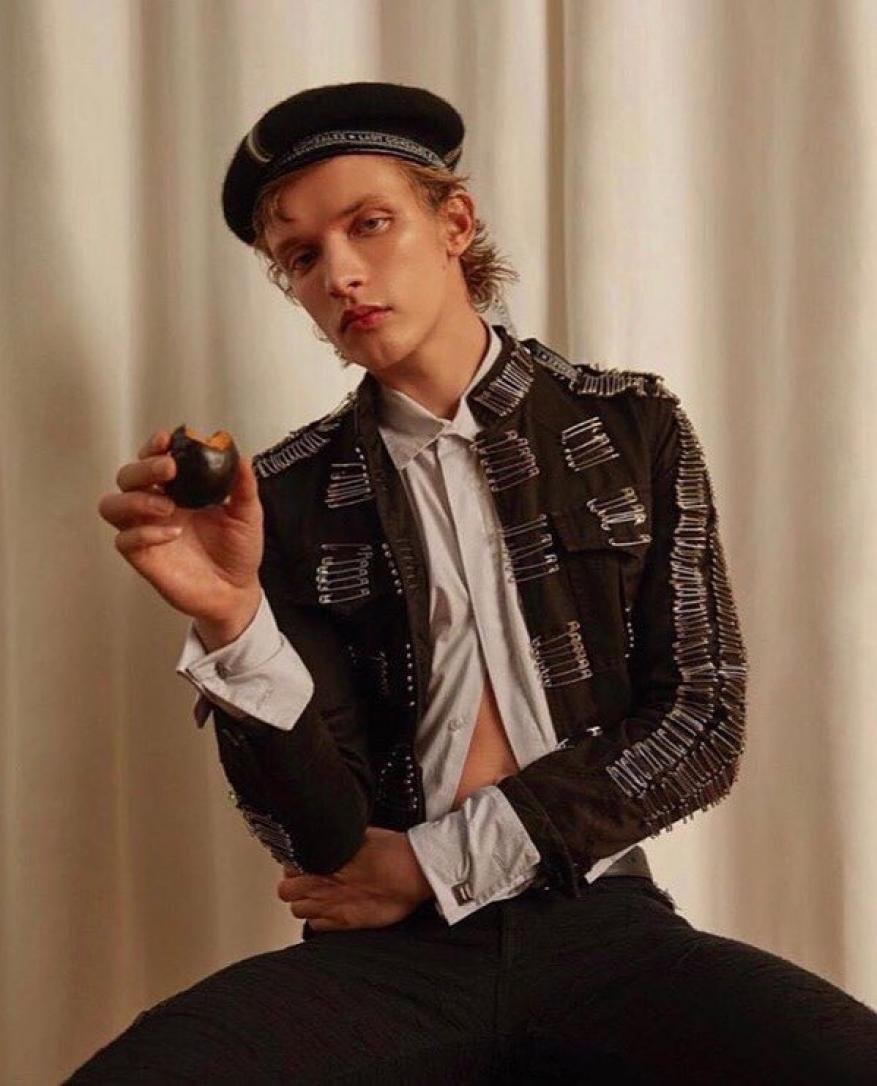 Young adult male with curly hair wears a beret and safety-pin-covered jacket, holding a black plum in a studio with cream background.