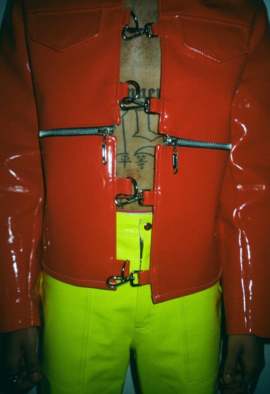 Person in red vinyl jacket with zipper and metal clasp details, paired with neon yellow pants against a plain backdrop.