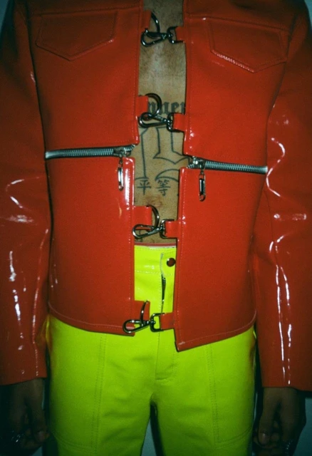 Person in red vinyl jacket with zipper and metal clasp details, paired with neon yellow pants against a plain backdrop.