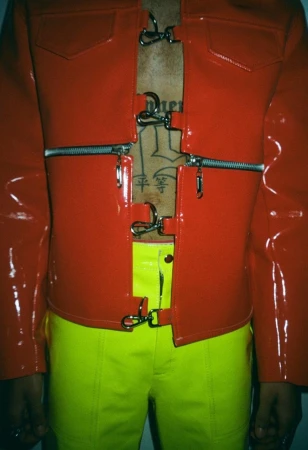 Person in red vinyl jacket with zipper and metal clasp details, paired with neon yellow pants against a plain backdrop.