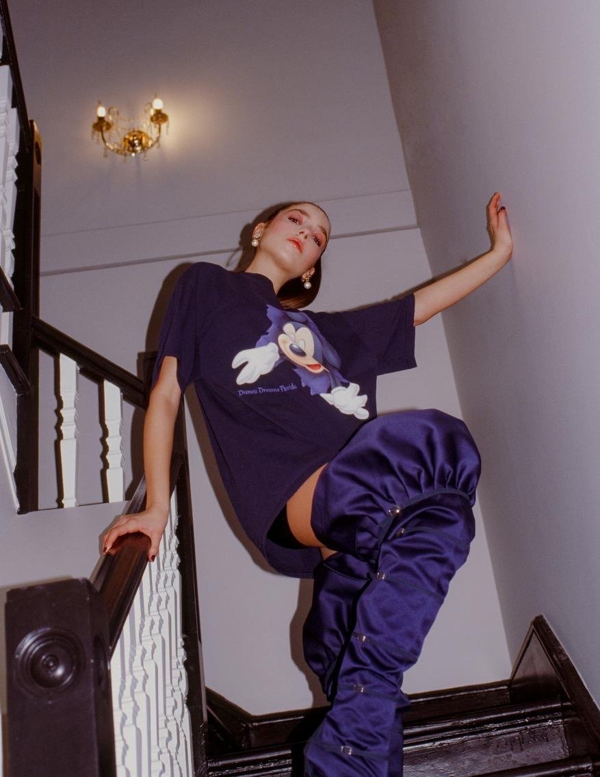A woman in a Disney T-shirt and tall blue boots poses on wooden stairs with a wall and light fixture visible in the background.
