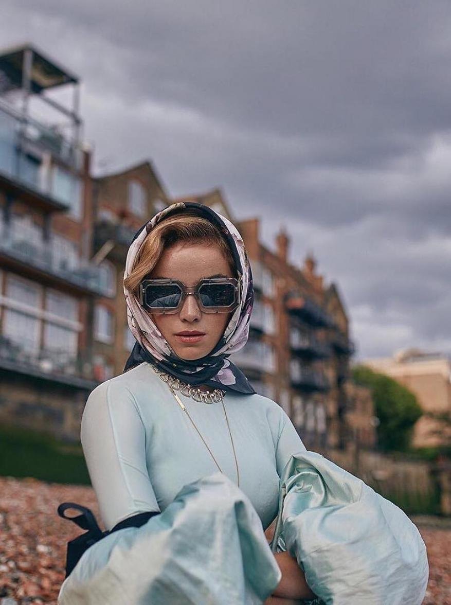 Woman in sunglasses and headscarf posing outdoors in front of brick buildings under an overcast sky, fashion shot, medium angle.
