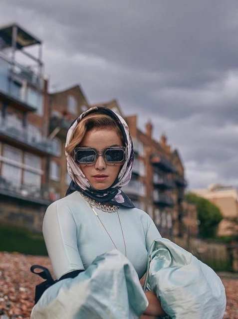Woman in sunglasses and headscarf posing outdoors in front of brick buildings under an overcast sky, fashion shot, medium angle.