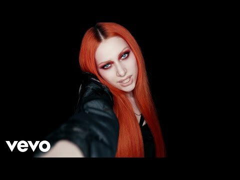 An adult female with red hair is shown in a medium shot against a black background, wearing a black jacket and vivid makeup.