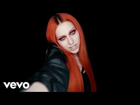 An adult female with red hair is shown in a medium shot against a black background, wearing a black jacket and vivid makeup.