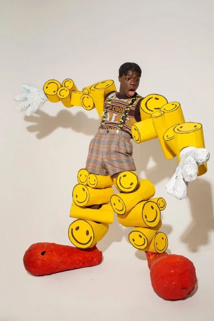 A young person in a brown plaid outfit with suspenders and yellow smiley face tubes posing in front of a white background.