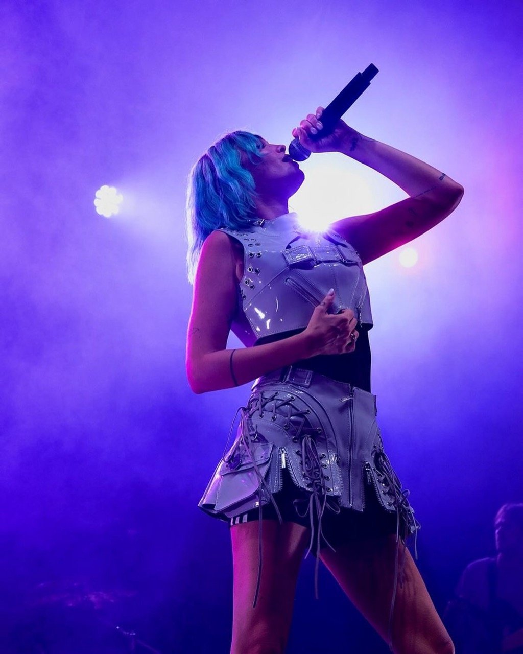 A singer with blue hair performs on stage, holding a microphone. The stage is lit with purple and white lights during the concert.