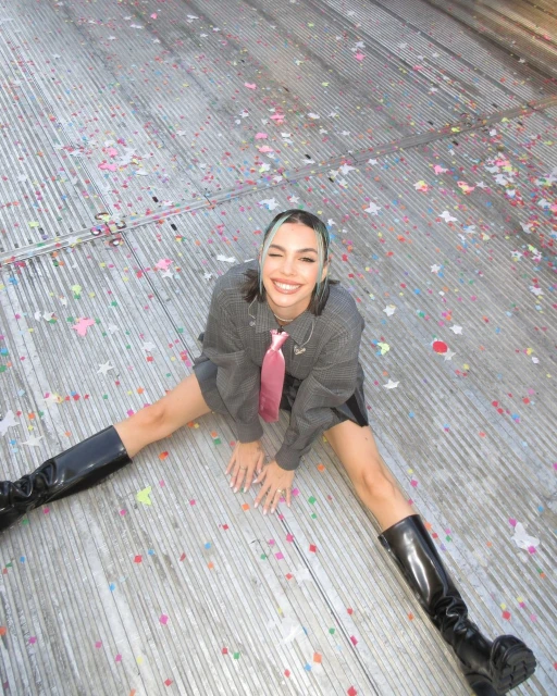 A smiling woman with a pink tie and black boots sits on a confetti-covered surface. Overhead shot focuses on her pose and confident expression.