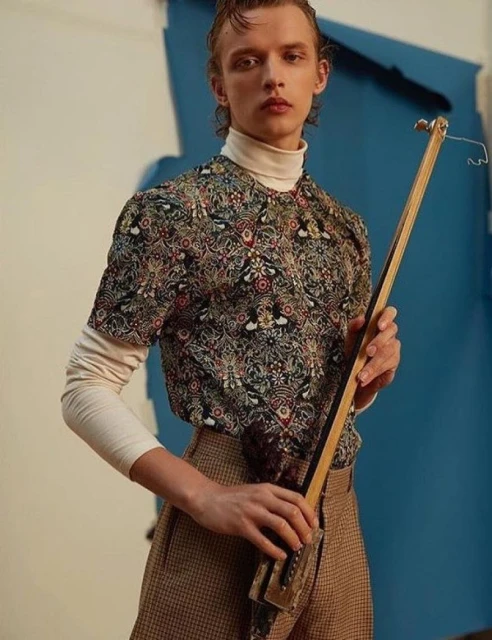Male fashion model poses in studio with ornate shirt, brown pants, and holding an unusual handcrafted wooden stringed instrument.
