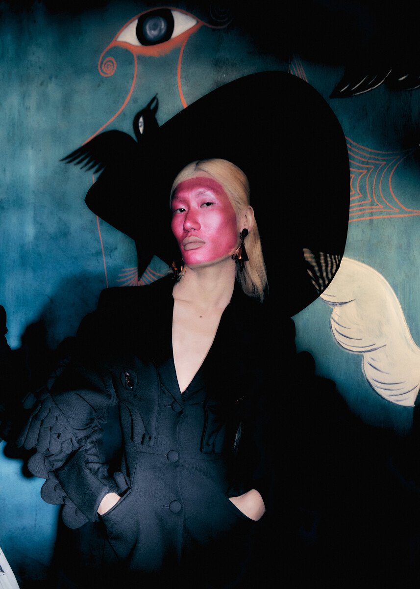Person poses in a black blazer and a wide-brimmed hat in front of a backdrop featuring an eye, a bird, and a cobweb.