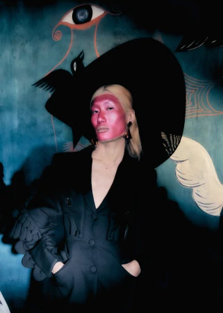 Person poses in a black blazer and a wide-brimmed hat in front of a backdrop featuring an eye, a bird, and a cobweb.