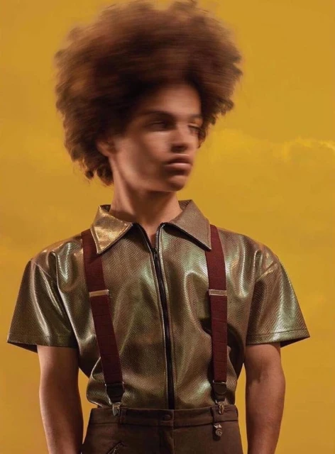 Young adult poses in gold shirt with suspenders against yellow background. Fashion editorial portrait showcases modern style.