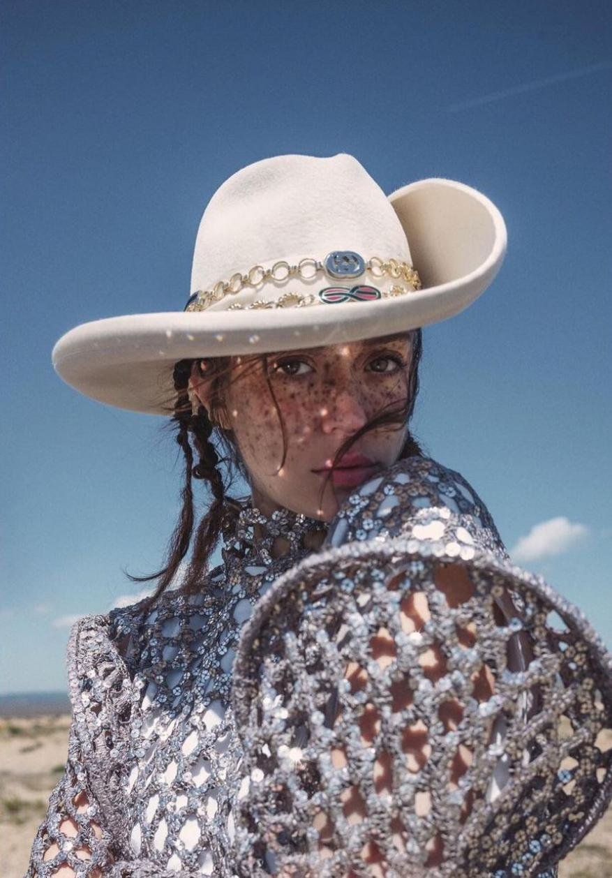A woman in a sequined top and cowboy hat poses in a desert landscape under a clear blue sky, captured in a close-up portrait.