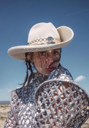 A woman in a sequined top and cowboy hat poses in a desert landscape under a clear blue sky, captured in a close-up portrait.