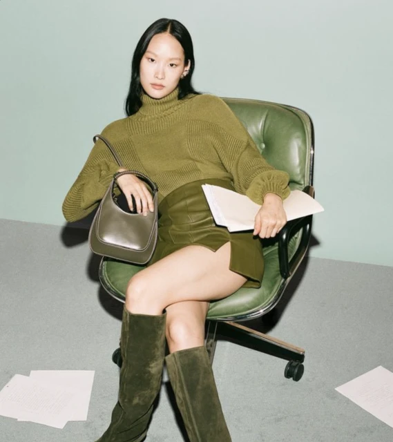 An Asian woman in an olive green sweater and faux leather skirt poses in an office chair with a purse and scattered papers.