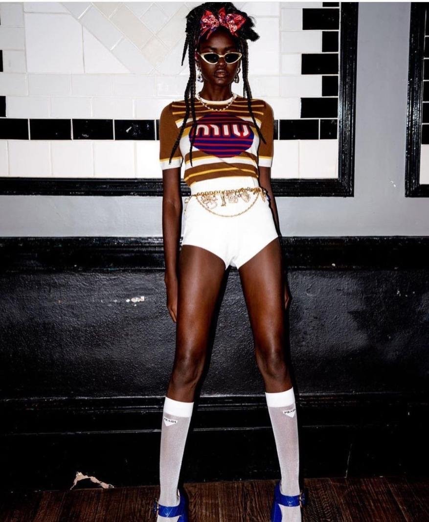 Dark-skinned adult female wearing a Miu Miu top, white shorts, and blue platform shoes poses in front of a white and black wall.