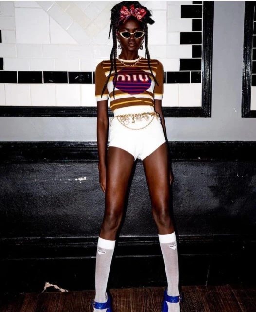 Dark-skinned adult female wearing a Miu Miu top, white shorts, and blue platform shoes poses in front of a white and black wall.