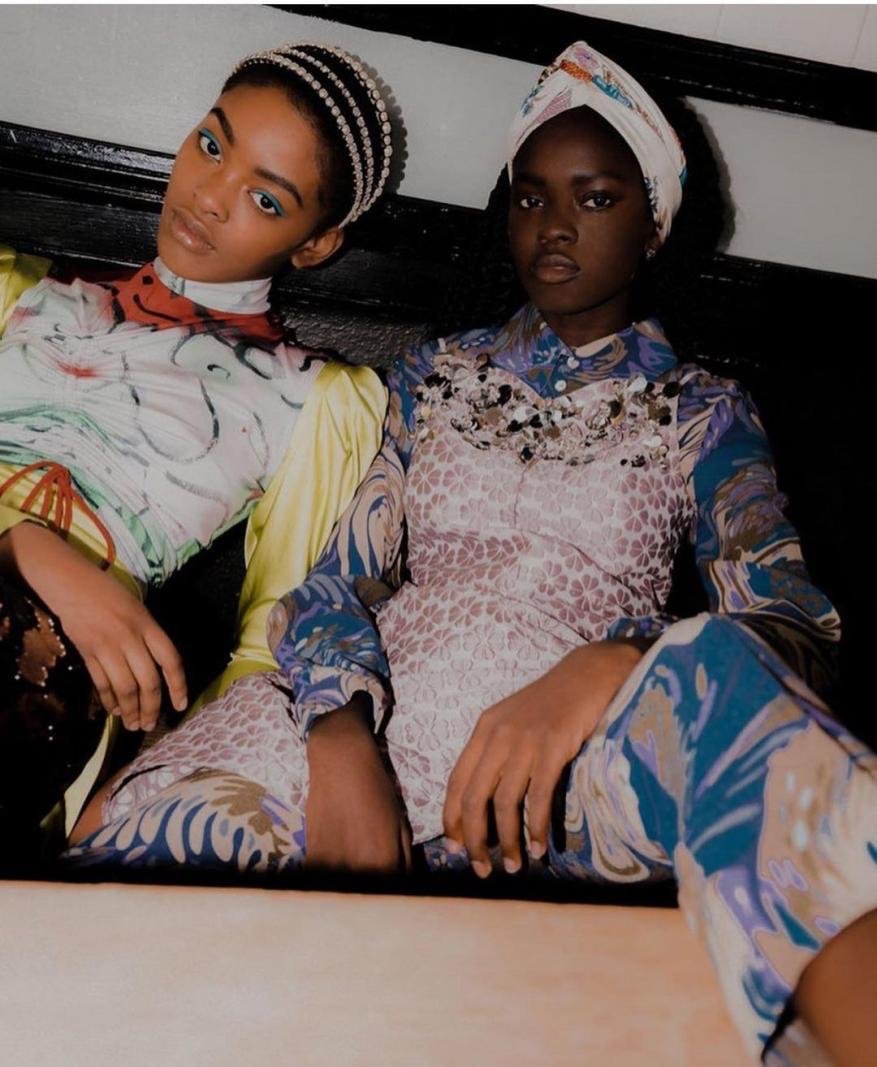 Two African American fashion models in patterned tops and head coverings pose indoors with direct gaze, showcasing style and beauty.