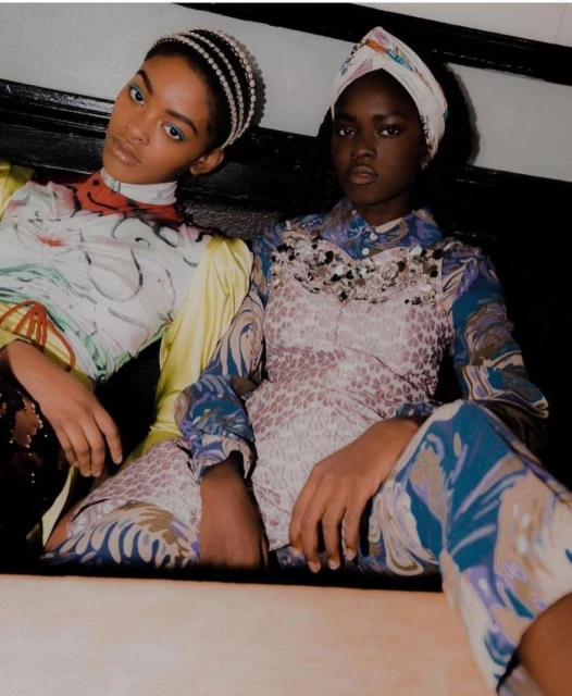 Two African American fashion models in patterned tops and head coverings pose indoors with direct gaze, showcasing style and beauty.