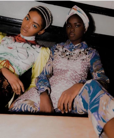 Two African American fashion models in patterned tops and head coverings pose indoors with direct gaze, showcasing style and beauty.