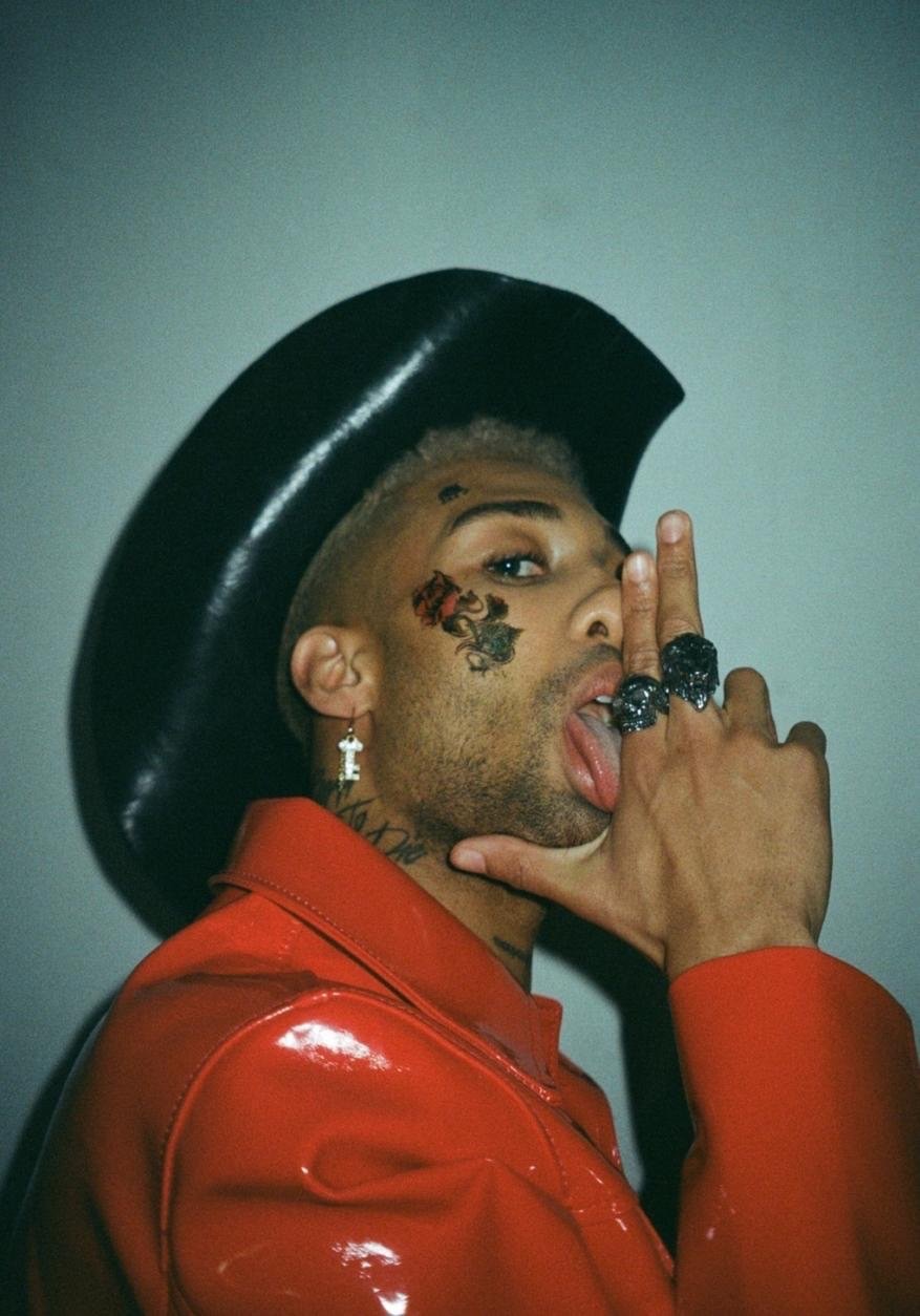Man poses with a cowboy hat and a red leather jacket, showing off tattoos and rings in a brightly lit indoor setting.