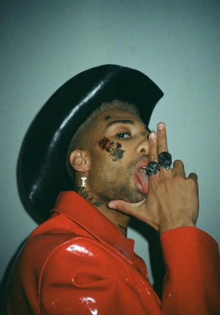 Man poses with a cowboy hat and a red leather jacket, showing off tattoos and rings in a brightly lit indoor setting.