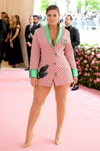 An adult woman poses on a pink carpet at an event, wearing a pink patterned blazer dress with green trim, tan heels, and printed tights.