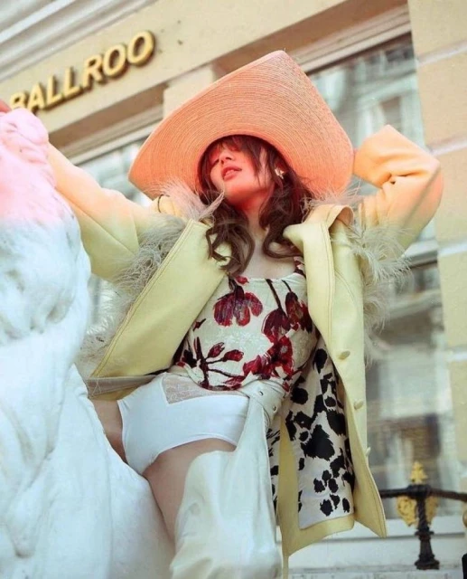 Woman in a large peach-colored straw hat and yellow coat poses next to a white swan sculpture outdoors, with a building visible behind her.