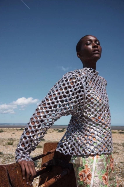 Fashion shot of a black woman in a silver mesh top, standing outdoors against a blue sky, wearing a floral skirt, and leaning on metal.