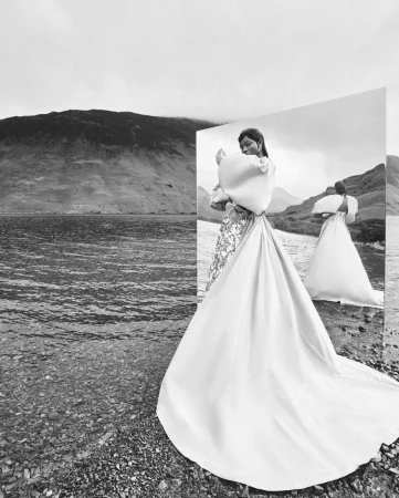Black and white editorial fashion portrait of a model in a voluminous gown standing next to a mirror on a rocky lake shore