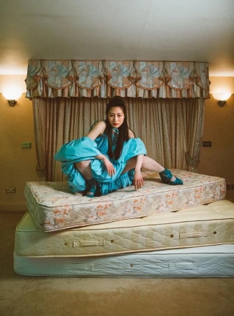 A model in a voluminous turquoise dress crouches atop a stack of mattresses in a traditional bedroom styled by Celia Arias