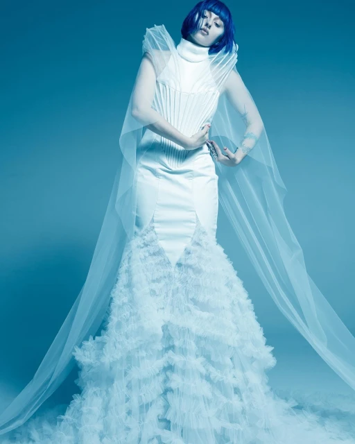 Woman with blue hair poses in a white wedding dress with a tulle train in a studio with a blue background, hands near chest.