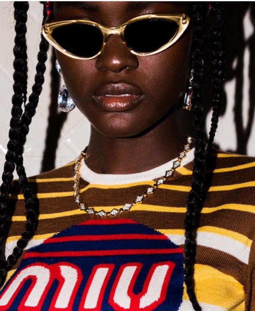 Close-up of a dark-skinned woman wearing gold cat-eye sunglasses, drop earrings, and a chain necklace in a fashion-style photo.