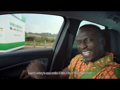 Man smiles inside car with car-buying business visible through window. Man wears bright orange patterned shirt in eye-level, medium shot.