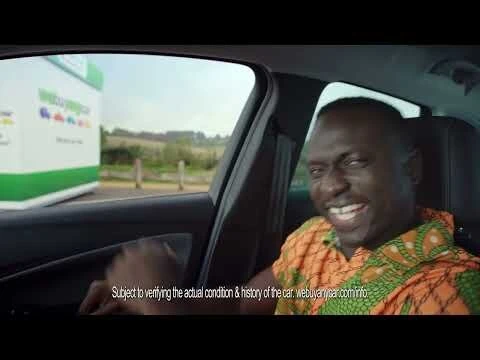 Man smiles inside car with car-buying business visible through window. Man wears bright orange patterned shirt in eye-level, medium shot.
