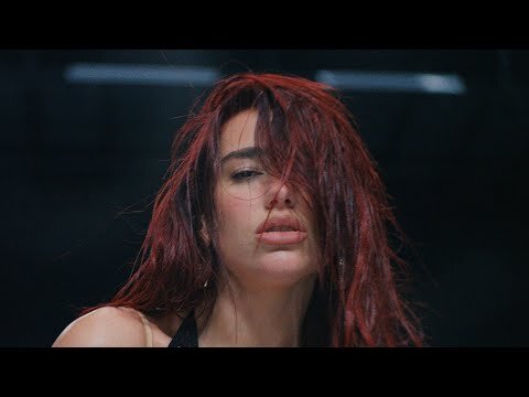 Portrait of Dua Lipa with vibrant red hair partially covering her face against a dark background