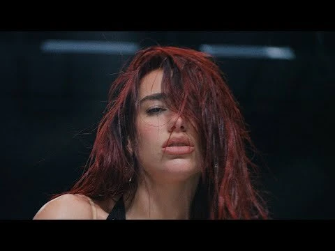 Portrait of Dua Lipa with vibrant red hair partially covering her face against a dark background